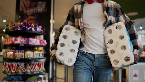 disparo en primer plano de un hombre con una camisa a cuadros con color de piel negra llevando enormes paquetes de toallas blancas desechables y caminando a lo largo de los mostradores con verduras y frutas en una tienda de comestibles moderna