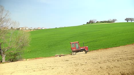 Pequeño-Tractor-Rojo-Que-Conduce-Cuesta-Arriba,-Equipo-De-Triple-Sistema-Rodante-Que-Prepara-La-Tierra-En-Una-Granja-Natural-Agrícola-Durante-Un-Día-Soleado-De-Primavera