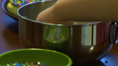 la mano de un niño sumerge una galleta casera en glaseado y luego en un tazón de chispas de colores