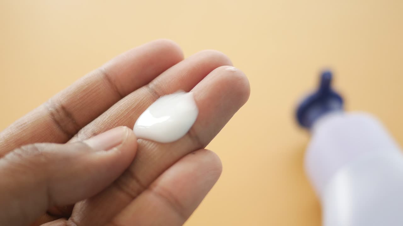 Close-up of a hand holding white lotion with a bottle of lotion in the background