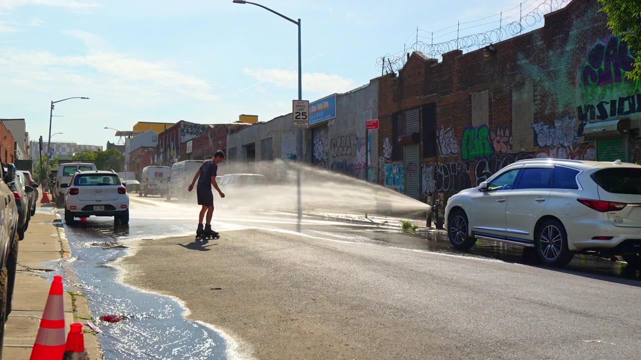 New York, USA, 5. juni 2026: Rulleskøjteløber passerer vandstråle fra brandhane på bygade. Biler, kegler og graffitivægge langs vejen
