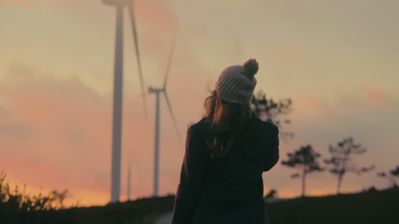 Gorgeous blonde woman in a gray coat, and beanie walks towards working wind turbines during late sunset in Portugal. Cinematic shot emphasizes green energy's significance and enjoying nature.