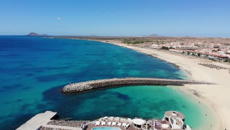 Aerial-View-Of-Idyllic-Blue-Ocean-Waters-With-Breakwater-Wall-Surrounding-Bikini-Beach-Club-At-Cape-Verde