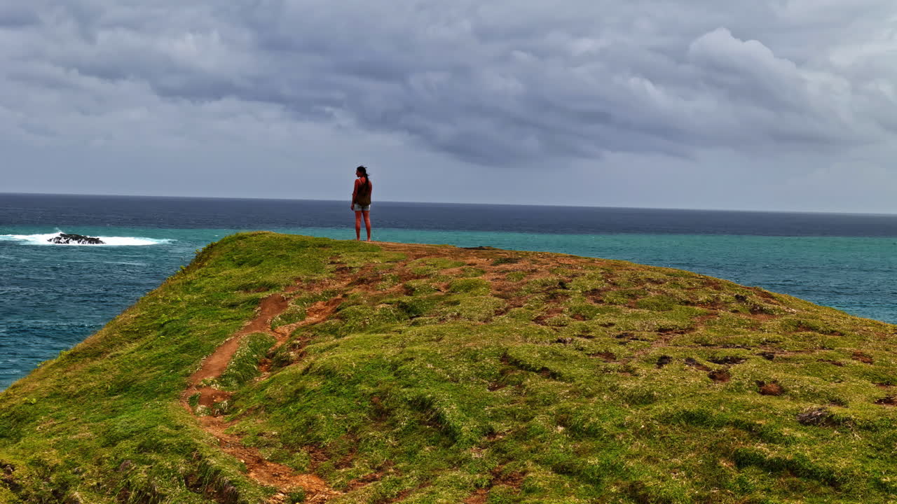 A pedestal shot rises from a lone woman standing on a grassy hill, unveiling the vast turquoise ocean and stormy sky in a dramatic Bali coastal scene