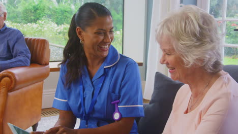 senior couple at home with woman talking to female nurse or care worker using digital tablet