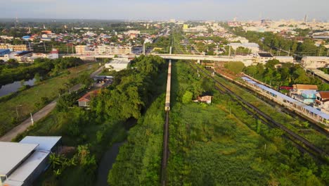 aerial shot: a long freight train passes under the highway bridge, enters the town in thailand, asia