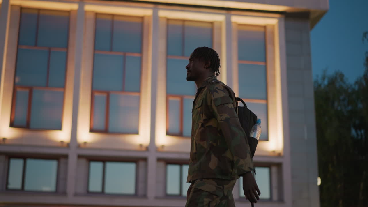 Evening Figure Against Illuminated Buildings, Shadowy Individual Moving Near Brightly Lit Urban Architecture, Silhouetted Person In Camouflage Coat Strolls Past Luminous Cityscape At Dusk