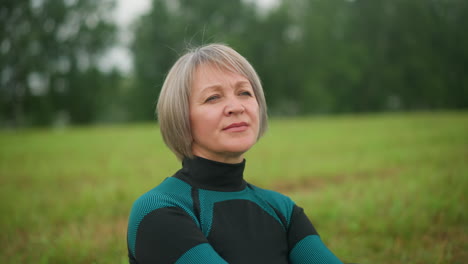 woman in green and black suit looking into distance thoughtfully with a warm smile in misty grassy field, surrounded by nature with blurred trees in background