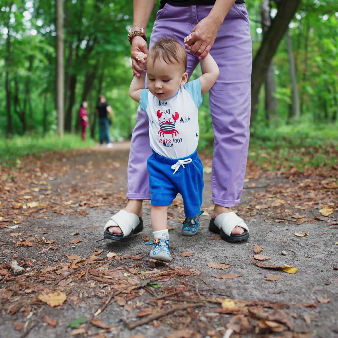 Teaching baby boy to walk. Mother is holding her baby by the hands and leading her child along the path in the park. Nature blurred backdrop