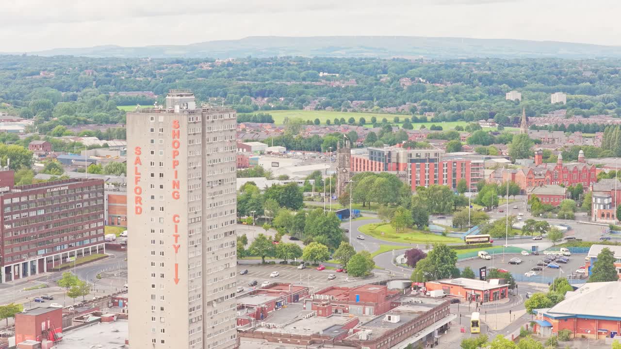 The Salford Shopping City tower rises above urban streets, car parks, and green spaces with distant hills on the horizon, captured in a slow descending aerial drone shot