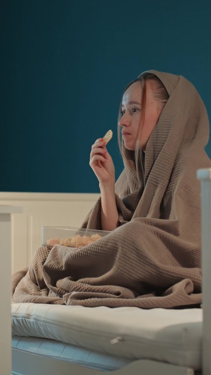 Una giovane donna siede sul letto coperta da una coperta marrone drappeggiata sulla testa. La donna mangia patatine ondulate da una ciotola di vetro trasparente. La scena è ripresa in primo piano, concentrandosi sulle azioni del soggetto.
