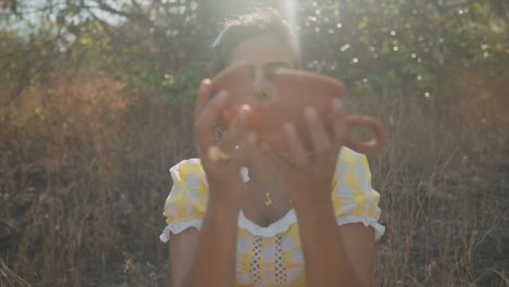 young woman standing in a field with dry grass attaching two broken pieces of a clay pot