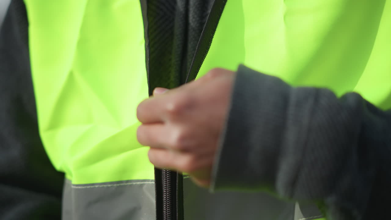 Close-up view of neon green reflective safety vest with black zipper partially open, showing detailed stitching and bright reflective strip under sunlight, emphasizing protective workwear design