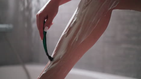 woman shaves leg skin hair with razor in bathroom closeup. graceful lady removes hair with blade in shower unit. woman with foam on leg and safe shaver