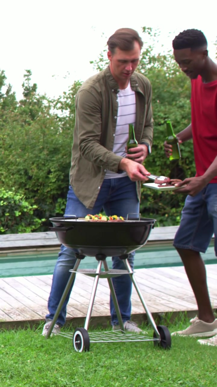 Video verticale: Uomo con maglietta color oliva che cucina al barbecue con bottiglia verde e pinze, mentre serve un amico che si avvicina.