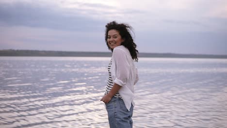 Smiling-young-brunette-caucasian-woman-in-white-shirt-and-jeans-holding-hands-in-pockets-feeling-free-standing-in-the-lake.-Outdoors,-morning-cloudy-sky