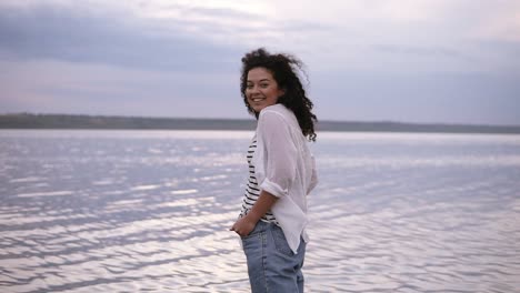 Sonriente-Joven-Morena-Caucásica-Con-Camisa-Blanca-Y-Jeans-Cogidos-De-La-Mano-En-Los-Bolsillos-Sintiéndose-Independiente-En-El-Lago.-Al-Aire-Libre,-Cielo-Nublado-Por-La-Mañana