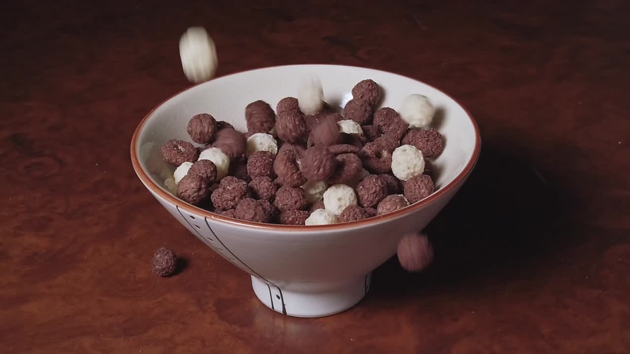 Chocolate crunchy cereals of different types fall into the bowl on a brown table. Slow motion. No milk
