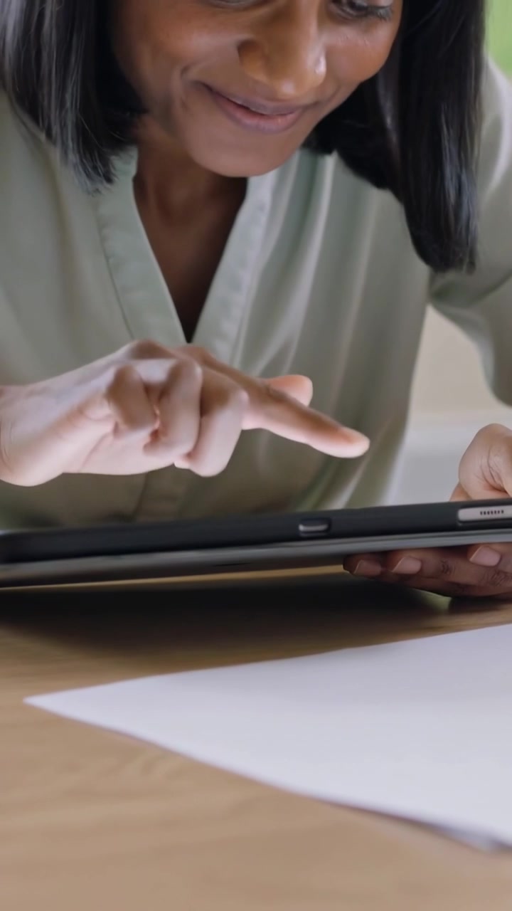 Video verticale: Donna con top verde che, dando un'occhiata allo schermo, tocca il tablet su un tavolo di legno con della carta