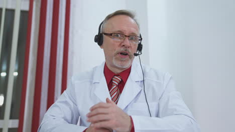 portrait of a doctor in headset. medical specialist giving a consultation online. professional male physician in white medical uniform speaks looking at camera.