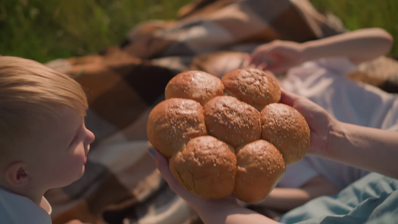 A woman s hand in a blue dress holds a cluster of freshly baked sesame seed bread rolls, gently placing them on a blanket in a grassy field. Her children are visible,slightly blurred in the background
