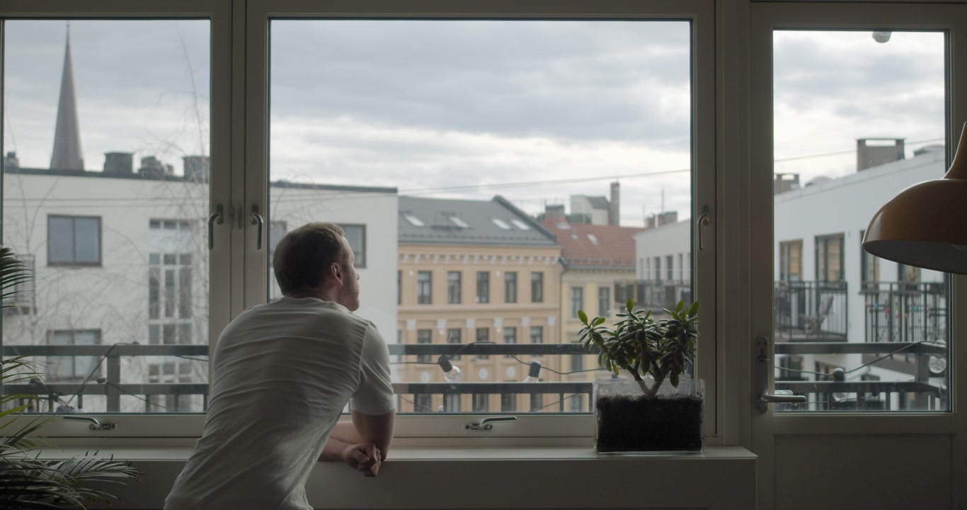 Plano cinematográfico amplo e estático de costas de um homem norueguês isolado sozinho em quarentena por coronavírus, olhando pela janela de um apartamento escuro com a cidade ao fundo, na parte central de Oslo, na Noruega.
