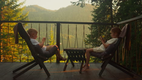 brother and sister send messages using modern mobile phones on balcony with breathtaking view. relaxed children have great time surrounded by untamed nature