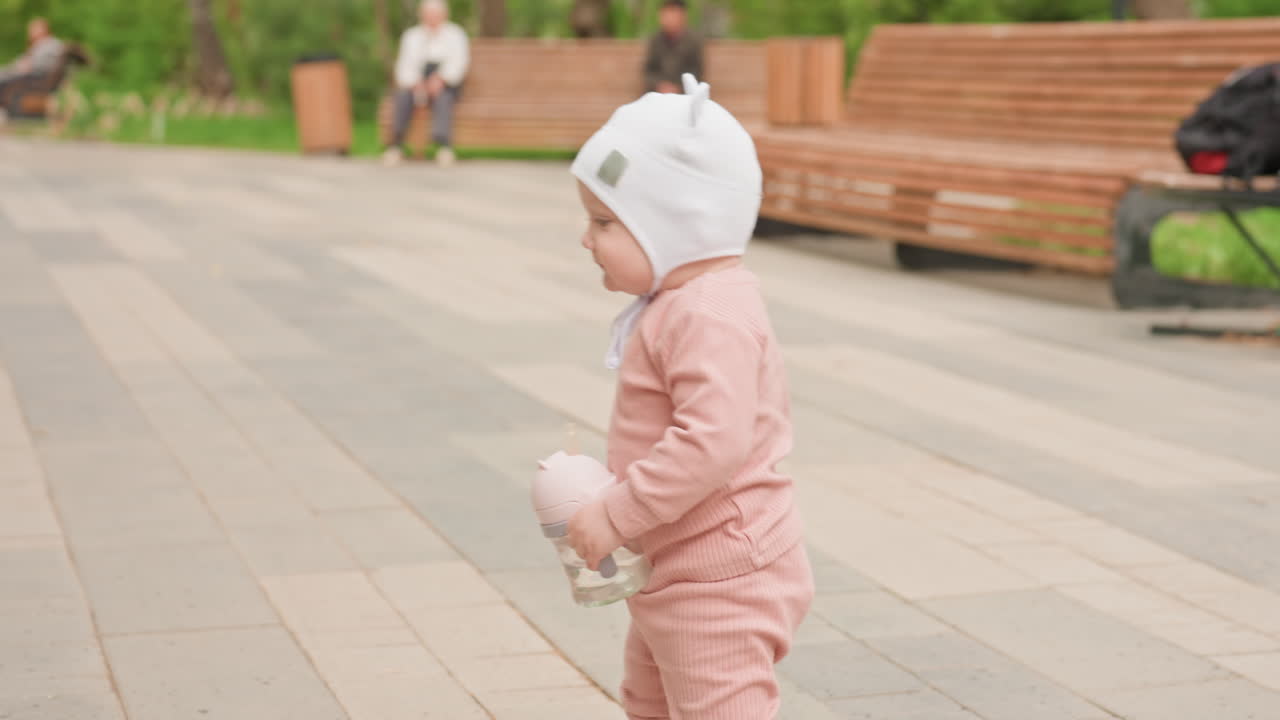 Small Toddler Exploring Near Guardian In Park, Young Child Takes Gentle Steps Past Parent On Paved Walkway, Little Toddler With Pink Outfit Moving Carefully Alongside Guardian In City Park Setting