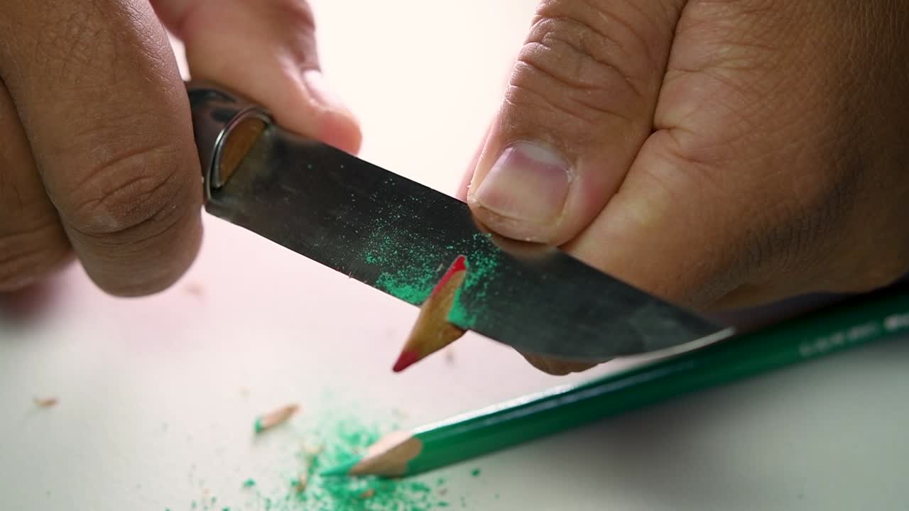 Footage of hands slowly sharpening a pencil and some coloured pencils with a sharp knife
