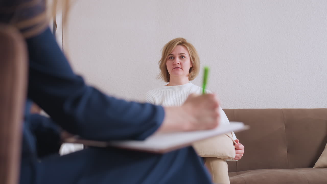 Conversation partner seated on sofa speaking expressively with therapist during session, captured in warm interior setting, with therapist taking notes attentively in foreground
