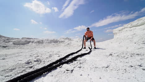 male athlete exercising outdoors around white hills at daytime. active physical workout. strong man in sunglasses has a workout with a rope on the ground.