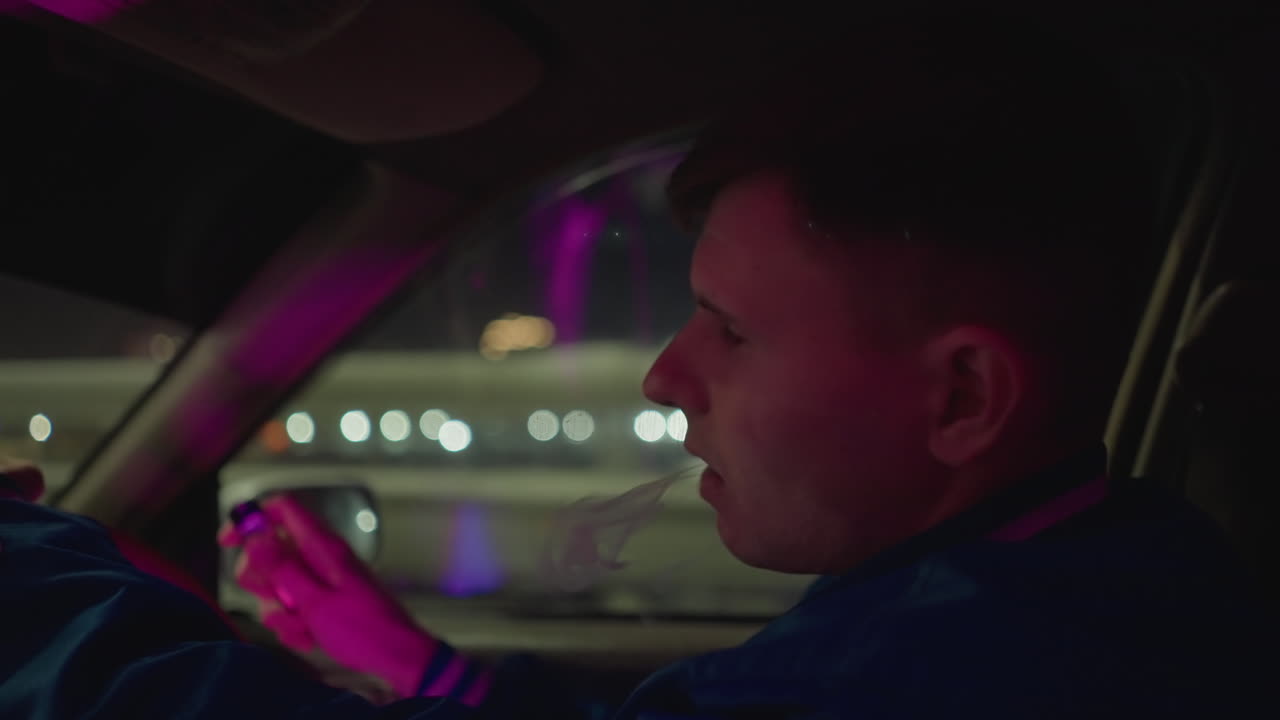 Interior de coche de noche: conductor vapeando con luces de neón. Primer plano del perfil de un joven agarrando el volante, llevándose el vape a la boca, mirando por el espejo y exhalando vapor contra las luces borrosas de la ciudad.