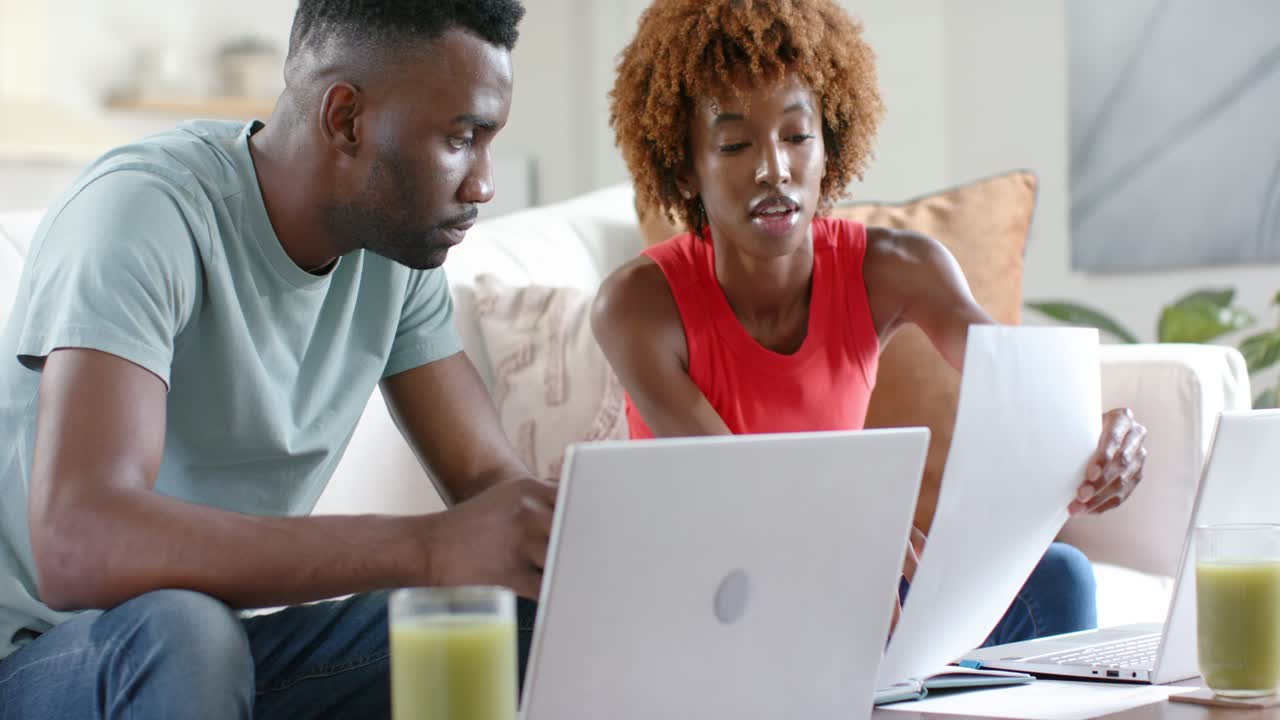 African American couple working, woman flipping papers urging man to type and check laptops on sofa