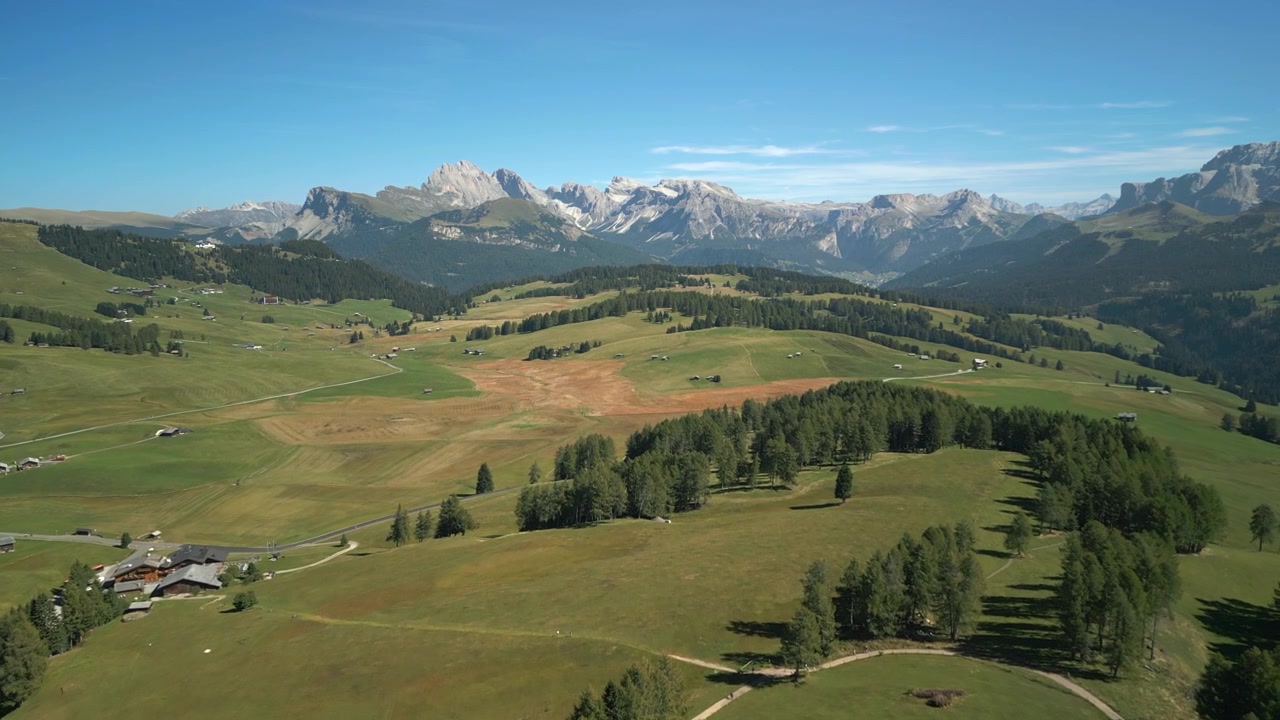 Ascesa aerea in avanti sopra baite alpine e ampi pascoli verdi, avvicinandosi a pareti rocciose delle Dolomiti italiane a mezzogiorno