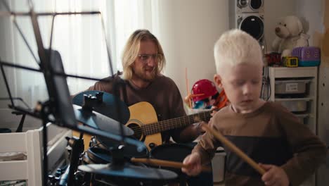 Un-Niño-Albino-De-Pelo-Blanco-Con-Un-Corte-De-Pelo-Corto-Y-Una-Chaqueta-Marrón-Toca-Una-Batería-Electrónica-Usando-Baquetas-Especiales-Junto-Con-Su-Padre,-Un-Hombre-Rubio-Con-Gafas-Y-Barba-Que-Toca-Una-Guitarra-Acústica-Con-Su-Hijo.