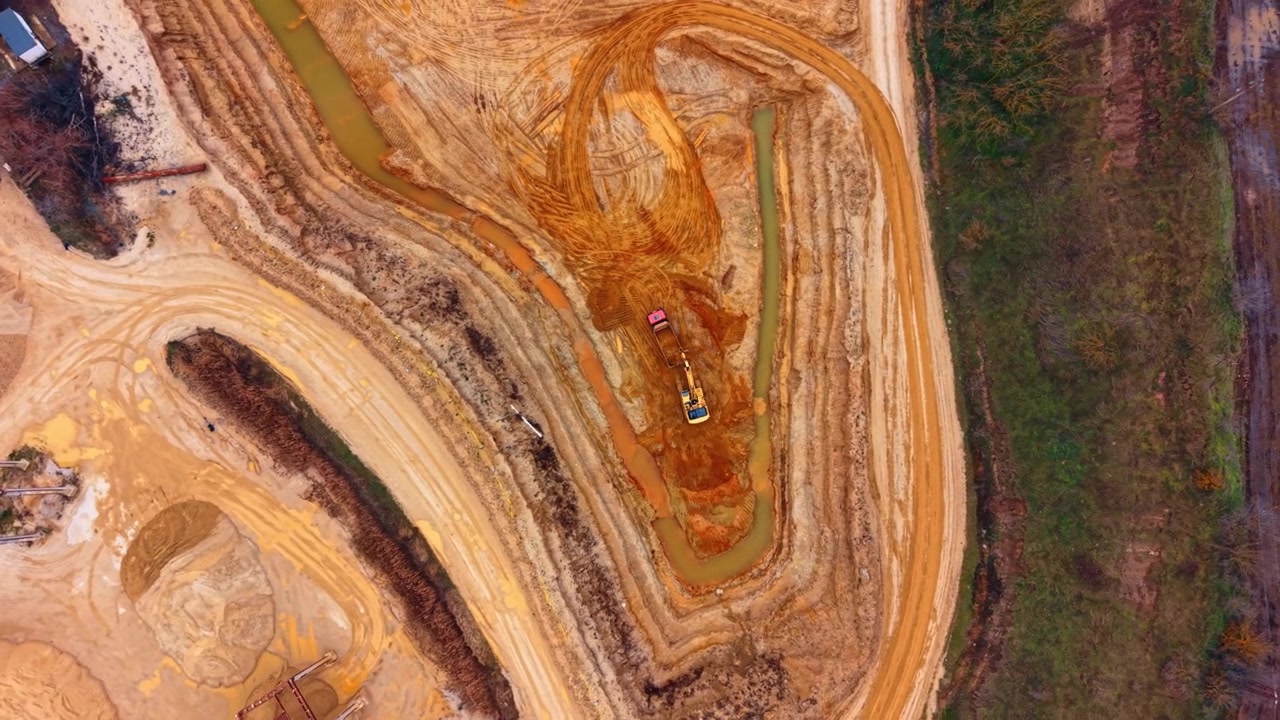 Excavator and truck working in layered clay quarry. Overhead drone view of two machines beside muddy water, tire marks and forest bordering the pit.