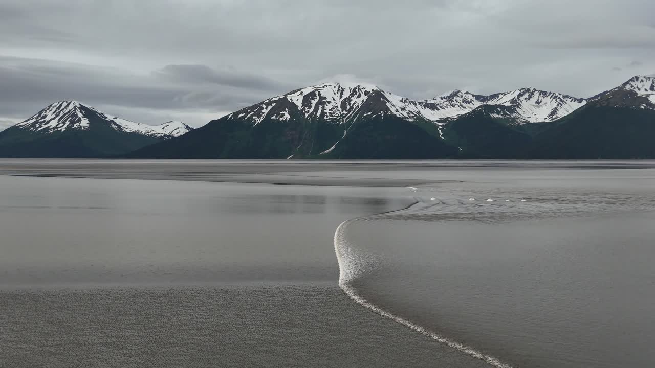 Drone Slowly Moving forward as Bore Tide Comes in on the Turnagain Arm in Alaska, Wave of water moving from right to left, Mountain with snow in background, Kenai Mountains