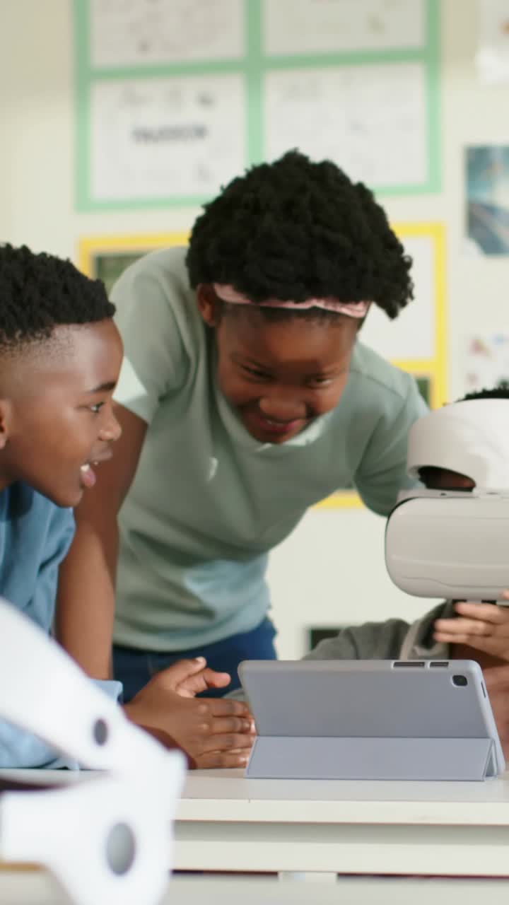 Vertical video: Guiding girl with pink band at classroom urging peer with white VR to reach tablet