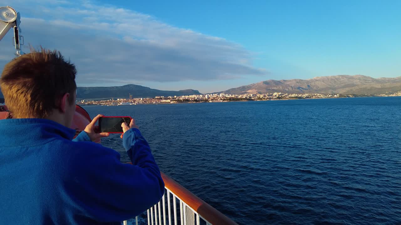 A tourist with a blue jacket and brown hair, during the corona pandemic, recording a video on his phone of coastline while the boat is sailing from Split to island Vis in Croatia, in the Adriatic Sea