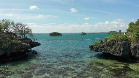 woman crossing wooden suspension bridge over clear tropical aqua waters between two rocky islands in 4k