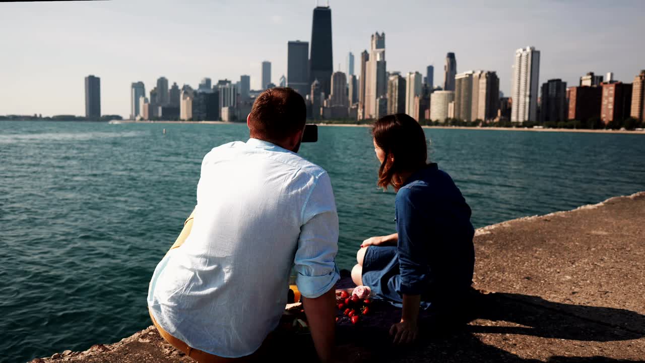 Back view of young couple having picnic on the shore of Michigan lake in Chicago, America. Man takes photo on smartphone