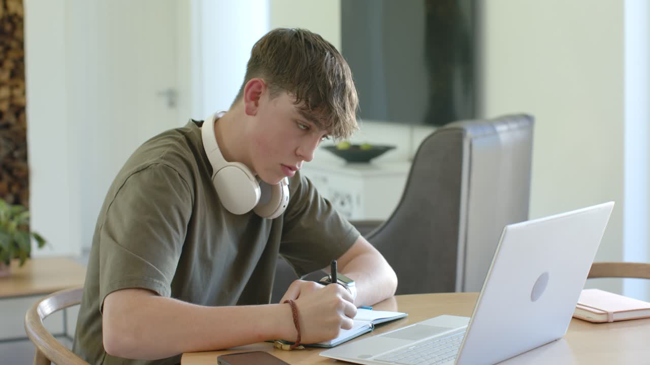 Tenåringsgutt studerer mens han svarer på en melding på laptopen og skriver i en liten notatbok ved spisebordet