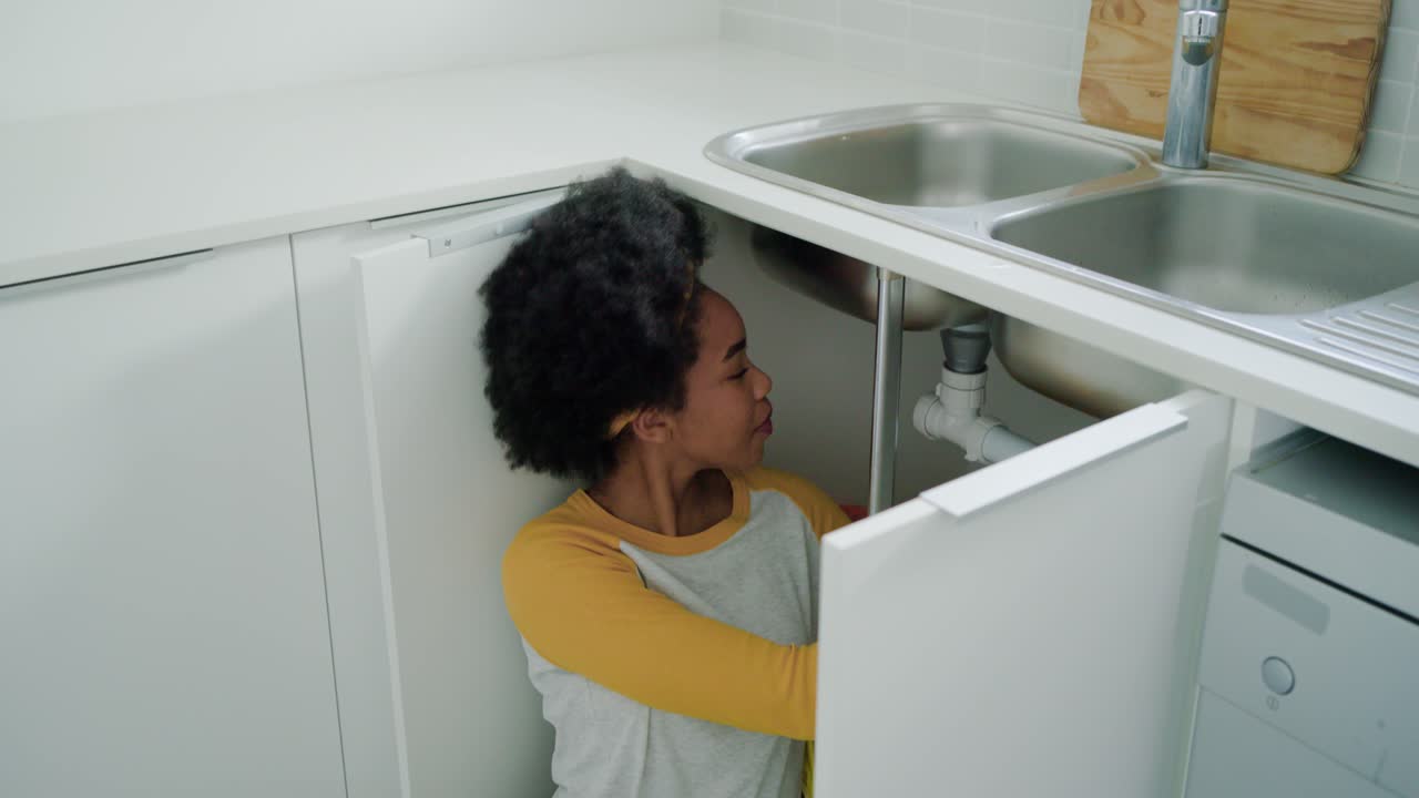 African American woman opening cabinet, checking, fixing pipes under kitchen sink using tools