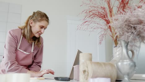 Young girl store owner of flowers and gifts works on laptop writes order smiling standing behind the counter of a bright stylish store