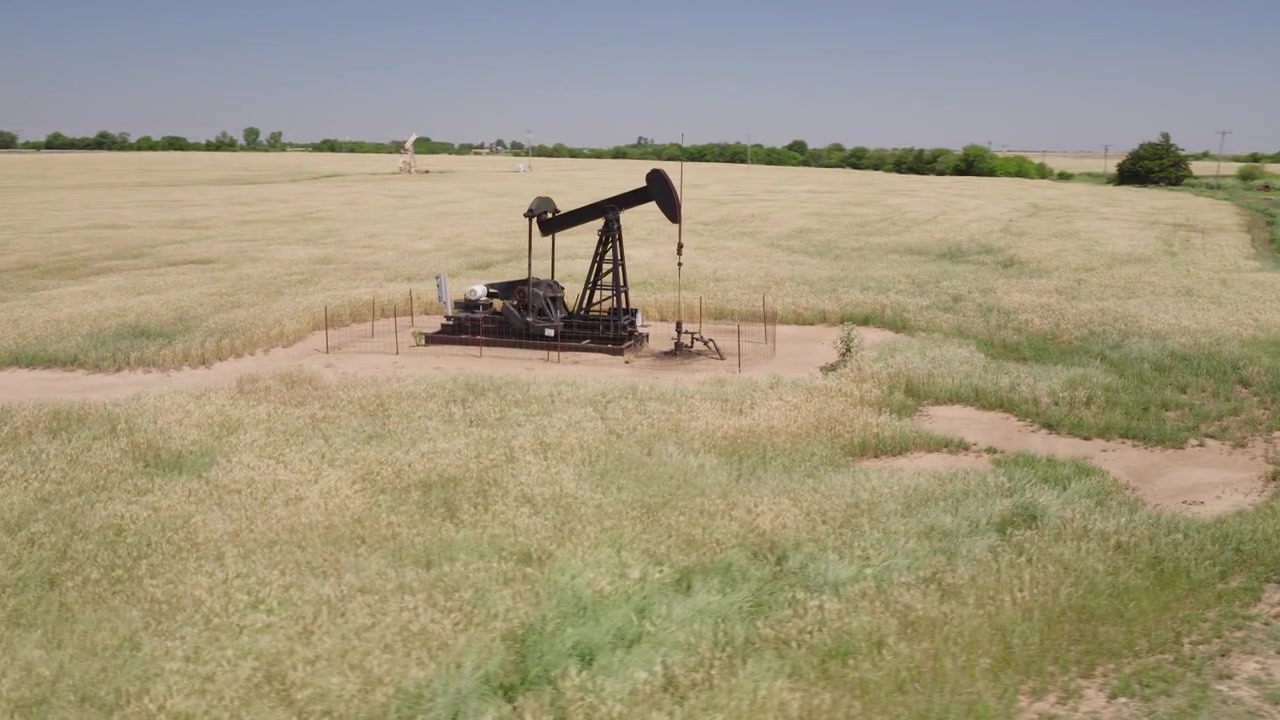 Drone 4K of a wheat field with two oil pump jacks and wind turbines in the background.