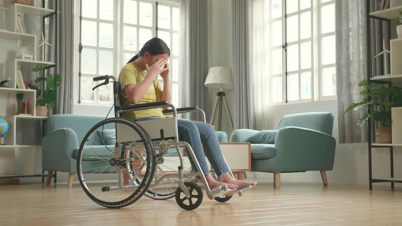 Disappointed Asian Kid Girl Sitting In A Wheelchair In Living Room At The House