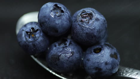 Nahaufnahme-Einer-Makroaufnahme-Von-Blaubeeren,-Die-Auf-Einem-Teelöffel-Spritzwasser-Auf-Einem-Dunklen-Hintergrund-Liegen,-Der-In-Zeitlupe-Erfasst-Wurde