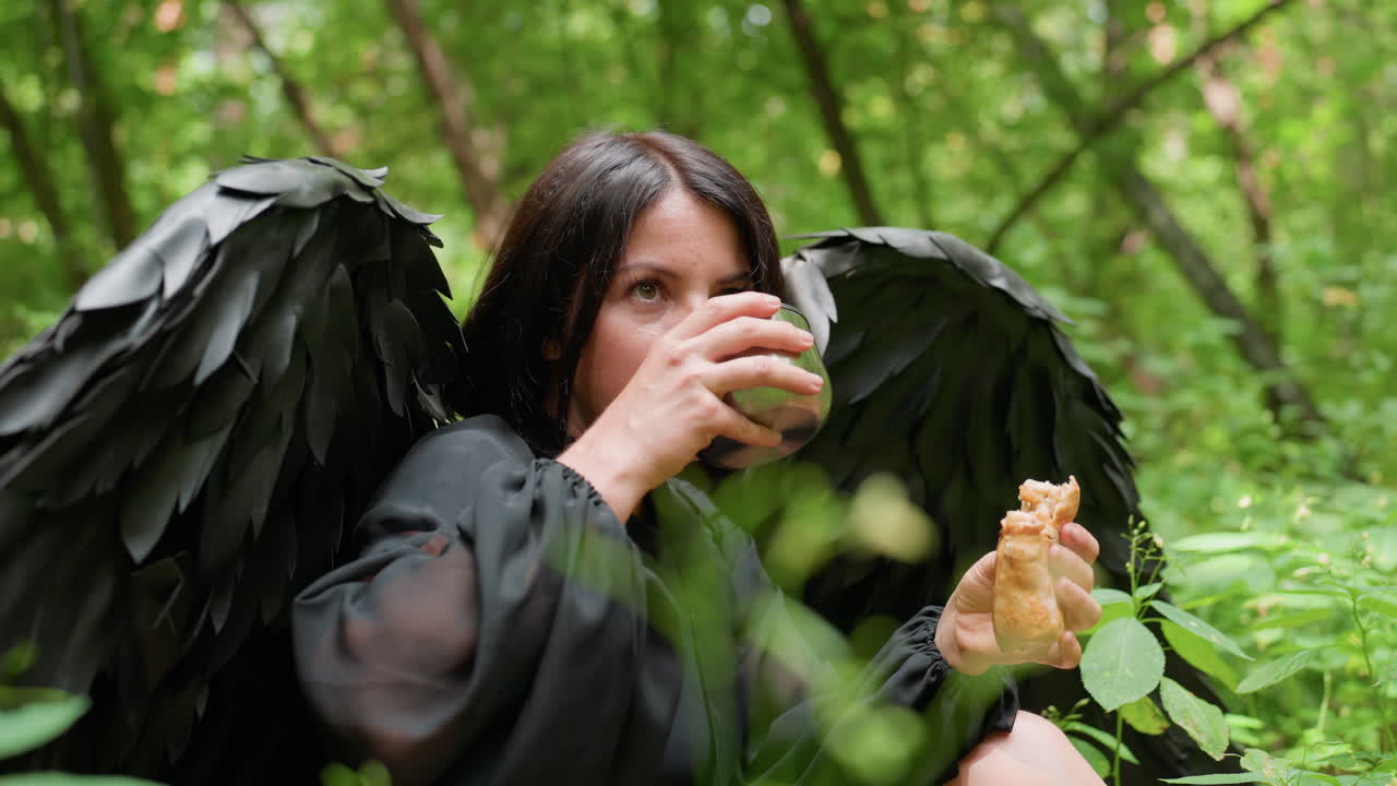 Young girl wearing black fairy costume with dark wings sits among lush forest greenery, savoring burger peacefully under soft daylight, combining mystical beauty