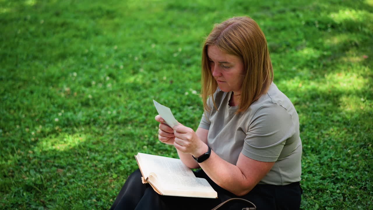 Young woman sitting on grass in peaceful park flipping through book discovering picture, gazing with curiosity under sunlight surrounded by trees, moment of reflection