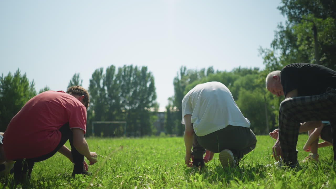 A family is seen from the back as they kneel on a sunny day, tying their shoelaces on a grassy field, in the distance, a goalpost is slightly blurred